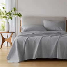 a bed with grey sheets and pillows in a white room next to a plant on a table