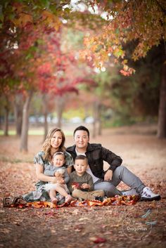 a family sitting on the ground in front of trees with leaves all around them and posing for a photo