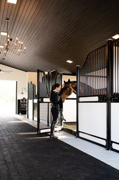 a person standing next to a brown horse in a stable with white walls and black railings