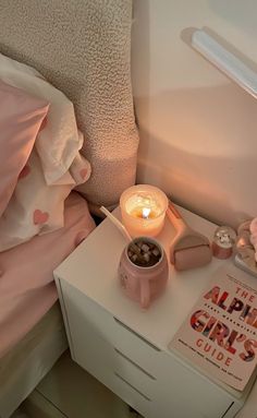 a night stand with a candle, cup and book on it next to a bed