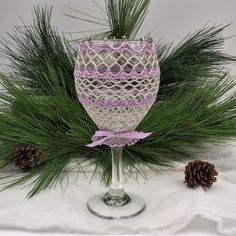 a wine glass decorated with pine cones and purple crochet sits on a white surface