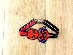 a red and black beaded bracelet with an orange fish on the clasp is laying on a wood floor