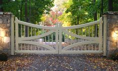 an open white gate in front of a stone wall and trees with leaves on the ground