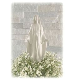 a white statue sitting on top of a lush green plant filled with flowers in front of a brick wall