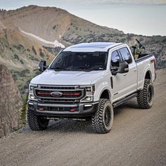 a white truck driving down a dirt road next to mountains and trees in the background