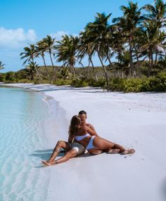 a man and woman sitting on the beach in front of palm trees, with water behind them