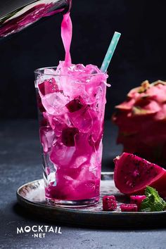 a pink drink being poured into a glass with ice and fruit on the table next to it