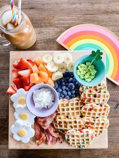 a wooden board topped with waffles, fruit and other foods next to a drink