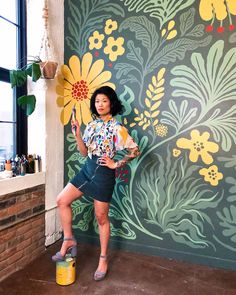 a woman standing in front of a wall with flowers on it and holding a yellow bucket