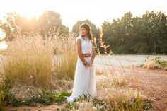 a woman in a white dress is standing by some tall grass and trees at sunset