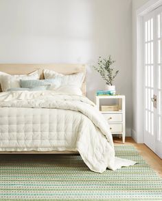 a bed with white sheets and pillows in a bedroom next to a window, rug and nightstand