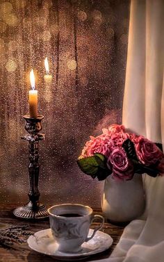 a cup of coffee next to a candle and some flowers on a table in front of a window