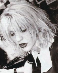 a woman with blonde hair wearing a tie and looking down at her cell phone while sitting in a chair