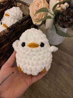 a hand holding a small white crocheted chicken in front of a basket with flowers