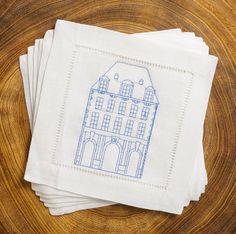 four white napkins with blue embroidered houses on them sitting on top of a wooden table