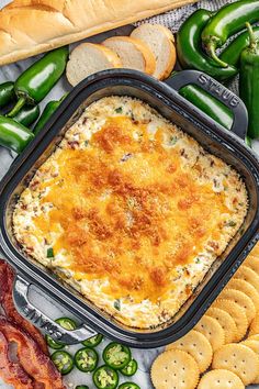 a casserole dish with cheese and bacon in it surrounded by crackers, green peppers, jalapenos and bread