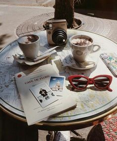 a table topped with two cups of coffee next to sunglasses and a camera on top of it