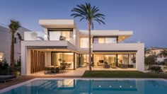 a large swimming pool in front of a modern house at dusk with palm trees and lawn furniture