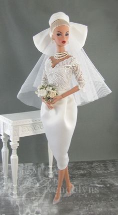 a barbie doll wearing a white wedding dress and veil with flowers in her hand standing next to a table