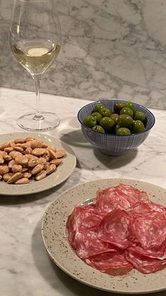 two plates filled with meat and olives next to a glass of wine on a marble table