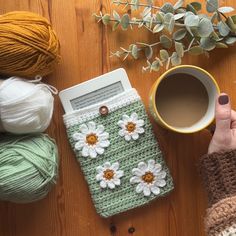 a woman holding a cup of coffee next to her cell phone and crocheted items