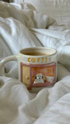 a coffee mug sitting on top of a bed covered in white sheets and blankets with a dog