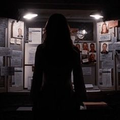 a woman standing in front of a wall full of papers and pictures at night with lights on