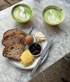 a plate with bread, butter and jam on it next to two cups of green tea