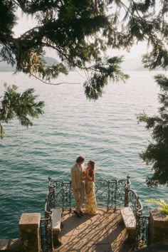 a man and woman standing next to each other on a pier near the water with trees