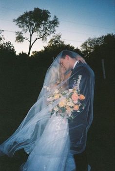 a bride and groom standing under a veil in the grass at sunset with trees behind them