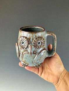 a hand holding a ceramic owl mug in it's left hand, against a gray background