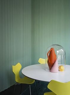 a white table with yellow chairs and a glass vase on top of it in front of a green wall