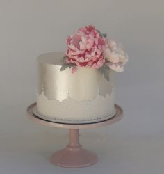 a white and silver cake with pink flowers on top, sitting on a stand in front of a gray wall