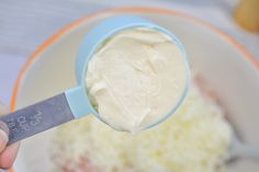 a hand holding a spoon with cream in it and some other ingredients behind the bowl