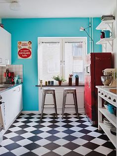 a black and white checkered floor in a small kitchen with blue walls, an old fashioned refrigerator and two stools