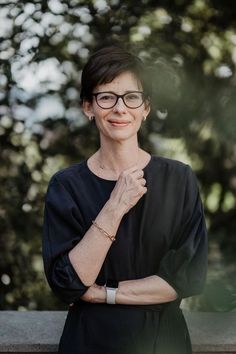 a woman wearing glasses standing with her arms crossed and looking off to the side in front of trees