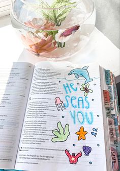 an open book on a table next to a vase with flowers and plants in it