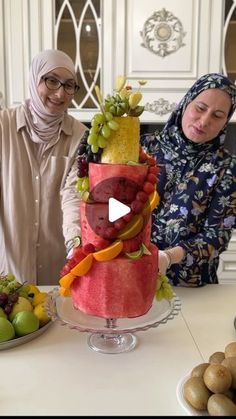 two women in hijab standing next to a cake with fruit on it and another woman behind the cake