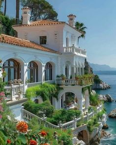 a house on the cliff overlooking the ocean with flowers and greenery growing all around it