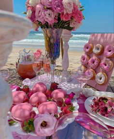 a table set up with donuts and pink flowers on it, near the beach