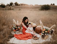 a woman in an orange dress holding a baby sitting on a blanket with flowers around her