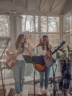 two young women are playing music together in front of a large window, while another woman is holding a guitar