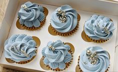 six cupcakes with blue frosting and pearls in a white box on a table