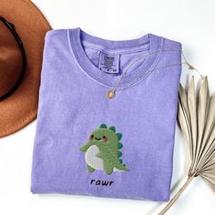 a purple t - shirt with a green dinosaur embroidered on it next to a brown hat