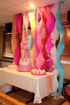 a table topped with cake and cupcakes next to colorful streamers hanging from the ceiling