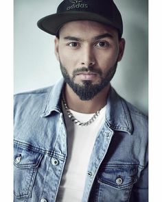 a man with a beard wearing a denim jacket and baseball cap is looking at the camera