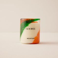 a close up of a candle on a white surface with the word goode written in green and orange