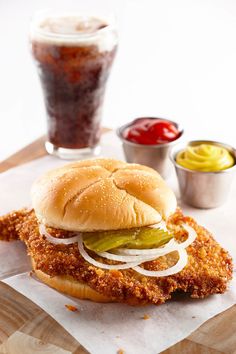 a chicken sandwich with pickles, onions and ketchup on a table next to condiments