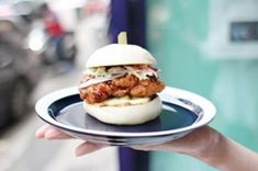 a person holding a plate with a chicken sandwich on it in front of a building