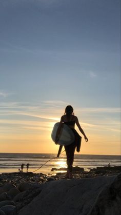 a woman holding a surfboard while standing on top of a rocky beach next to the ocean